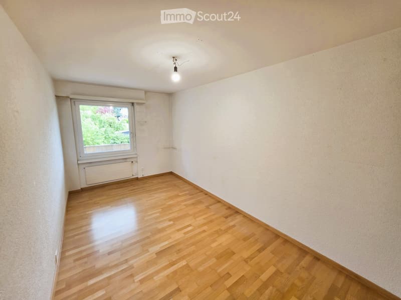 Empty room with wooden flooring, white walls, and a single window with a view of greenery outside.