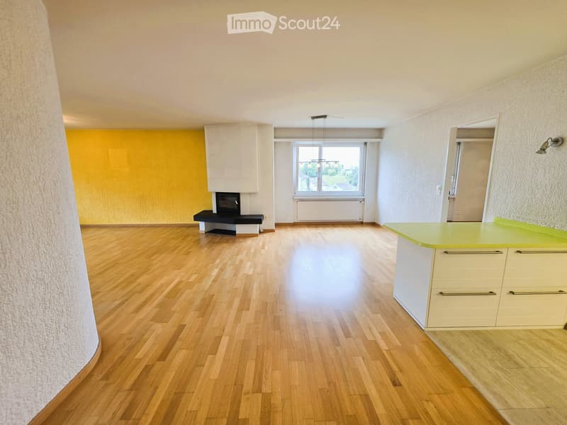 Bright, open room with wooden flooring, a small kitchen area with cabinets, a fireplace, and a window offering a view of the outdoors.