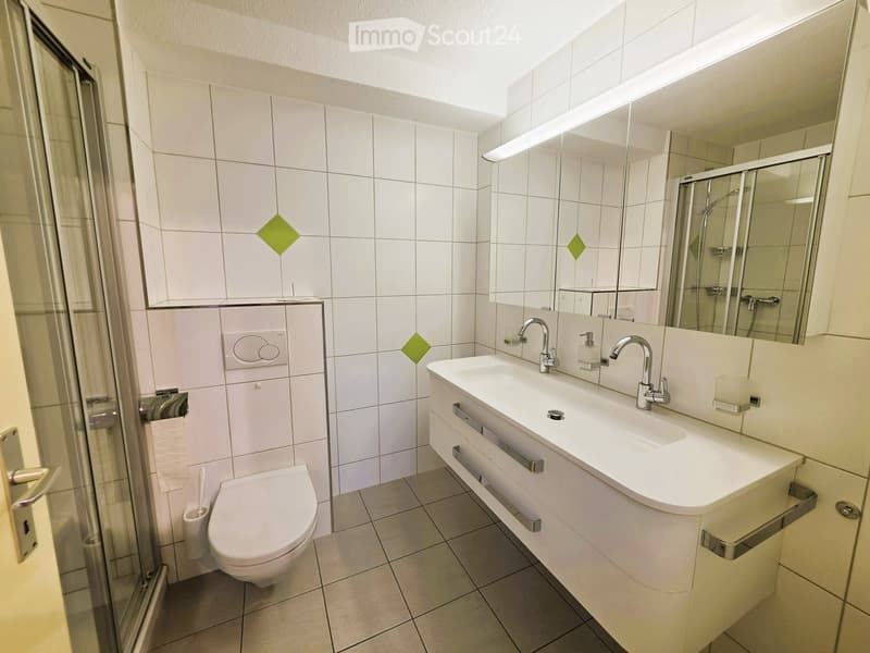 Modern bathroom with white tiles, a double sink, a toilet, a shower area, and a large mirror with built-in lighting.