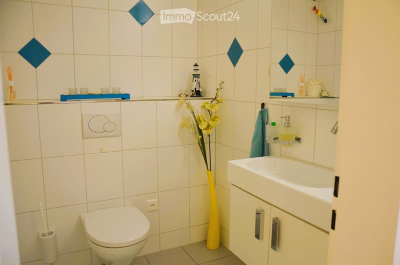 white tiles, white toilet, white sink, yellow vase with flowers, blue diamond tiles, white toilet paper holder, soap dispenser, towel rack