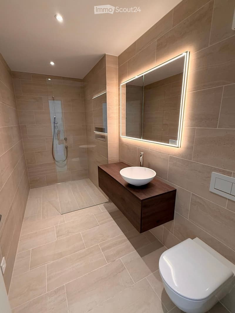 Modern bathroom with a shower, a sink, and a toilet. Beige tiles on walls and floor, a wooden vanity with a sink, and a large mirror with lit edges.