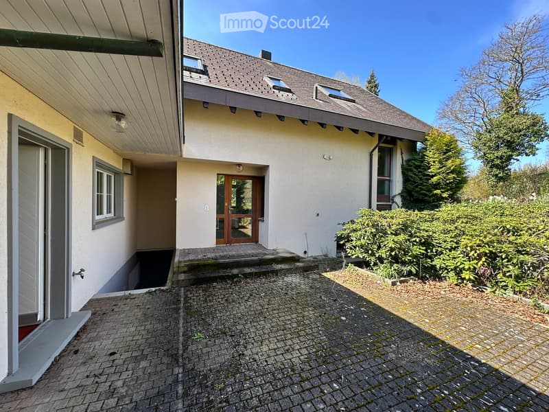 Raised ground floor, white exterior, brown roof, wooden door, brick driveway, shrubs, trees