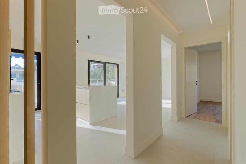 Empty hallway with white tiles, wooden floor in one room, kitchen with island, glass doors, and windows