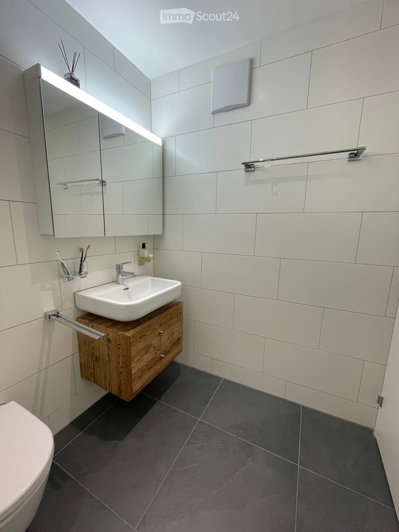 modern bathroom with white tiles, wooden cabinet, white sink, mirror, toilet, and towel rack