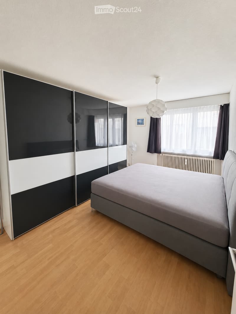 Bedroom with wooden floor, large bed, large wardrobe, white walls, large window with curtains, radiator, ceiling lamp