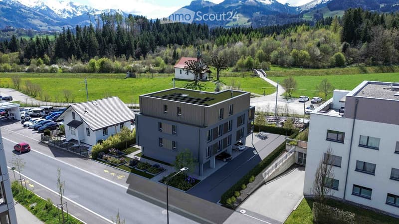 Aerial view of a modern apartment building with a parking lot, surrounded by greenery and mountains