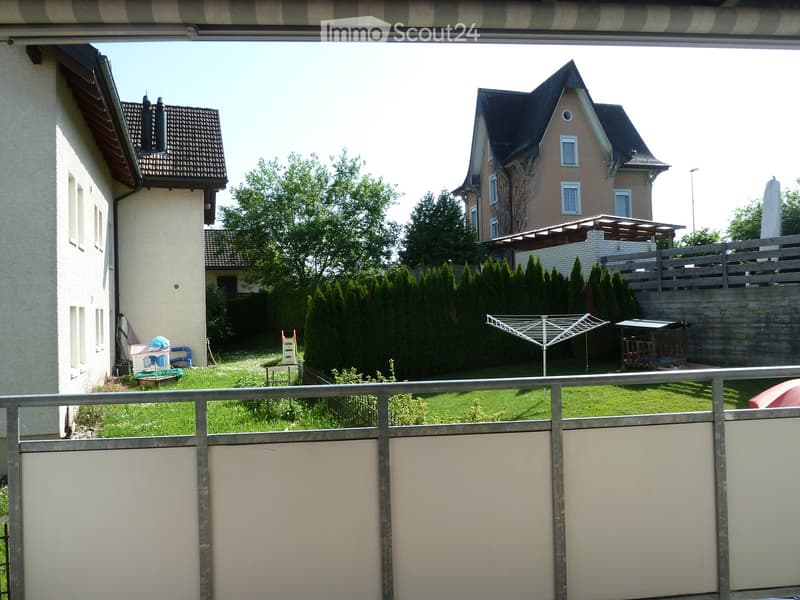 View from a balcony overlooking a well-maintained garden with a clothes drying rack, a small playhouse, and surrounding houses with pitched roofs and balconies.