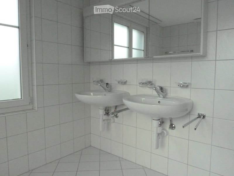 Clean bathroom with two white sinks, silver faucets, and a large mirror above them. The walls and floor are tiled in light gray.