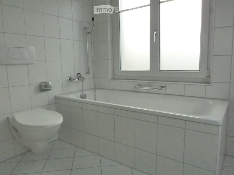 A clean, tiled bathroom with a bathtub, toilet, and a window above the tub. The walls and floor are covered in light gray tiles, and the fixtures are modern and functional.