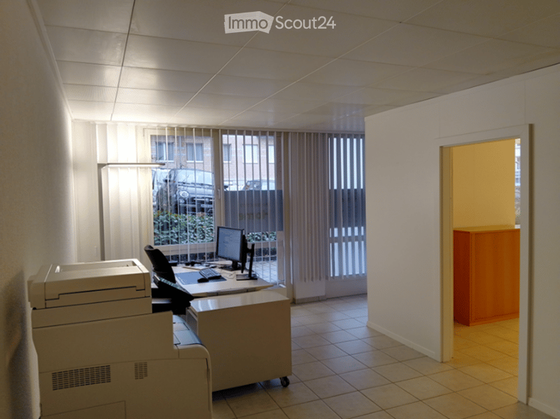 An empty office with a desk, computer, printer, and a cabinet. The room has white walls and tiled flooring. There is a window with blinds and a view of a car parked outside.