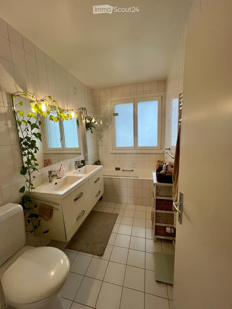 Bathroom with white tiles, double sink, bathtub, toilet, mirror, and a small shelf with toiletries.