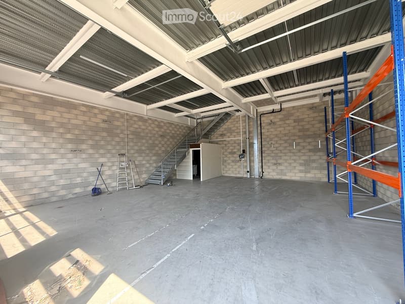 Empty warehouse space with concrete floor, exposed metal ceiling, brick walls, and a staircase leading to an upper level. Scaffolding and ladders are present.