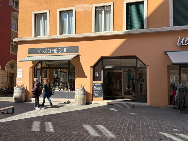 A two-story building with a wine shop on the ground floor, featuring a glass door, a blackboard with text, and wooden barrels outside. The building has several windows and a sign reading 'VINOTHEQUE' above the entrance.