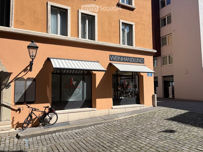 A building with a sign that reads 'Weinhandlung', two windows with awnings, a bicycle parked in front, and a lamp on the wall.
