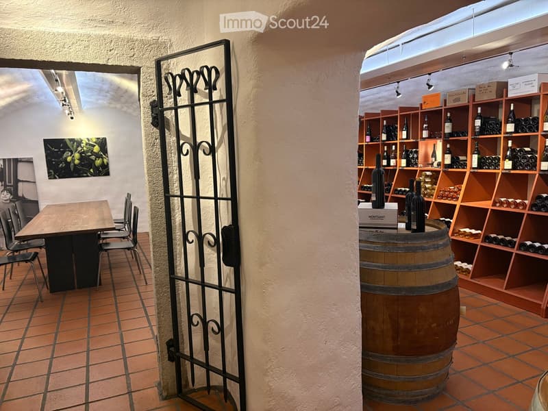 A room with a wooden table, chairs, a black gate, a barrel, shelves with wine bottles, and a painting on the wall.