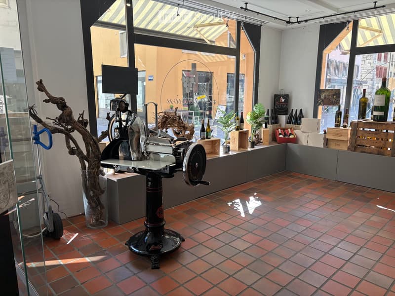 A room with a large window, a black machine, a glass vase with a tree branch, a trolley, and a shelf with wine bottles and boxes