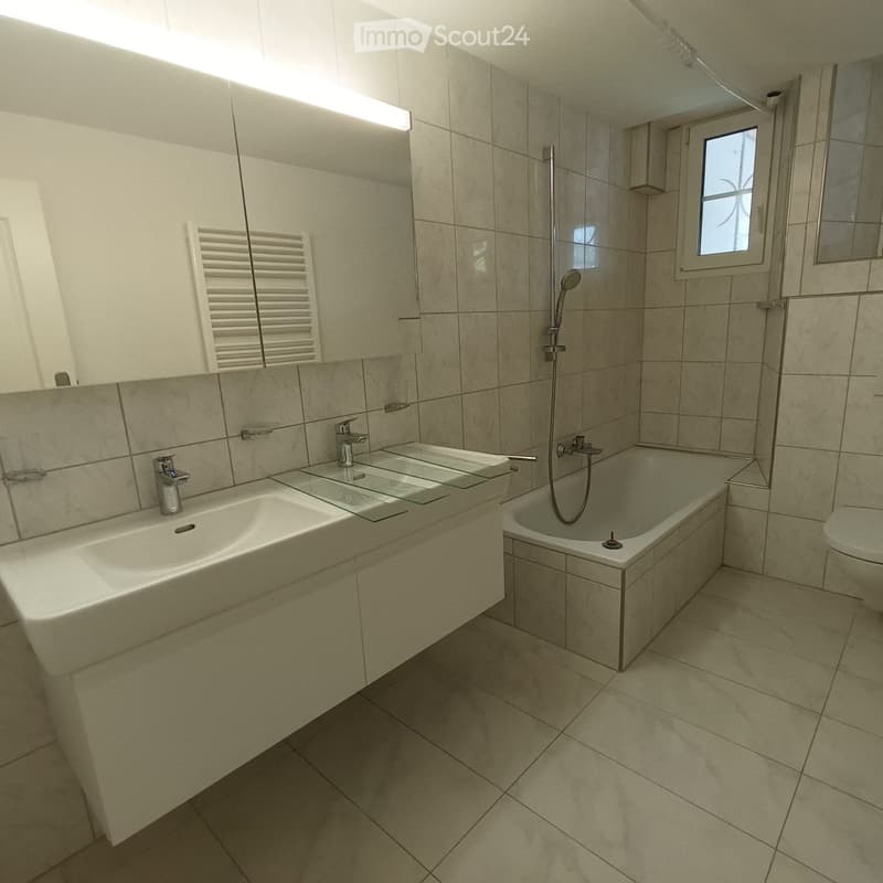 Modern bathroom with beige tiles, a double sink, bathtub, and a showerhead, illuminated by natural light from a small window.