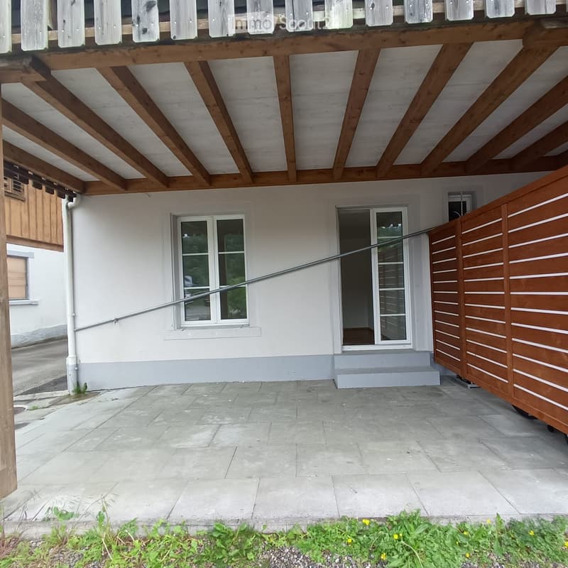 Outdoor patio area with stone tiles, wooden pergola, white house with windows, and small garden bed with plants and flowers.