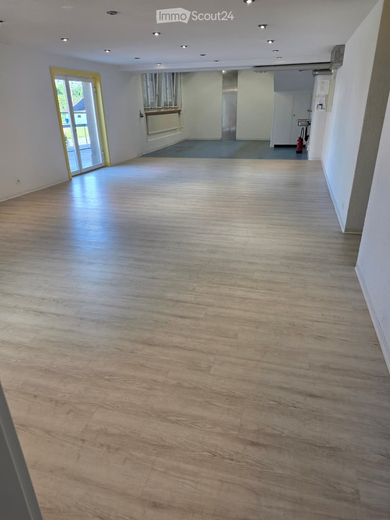 Empty room with light wooden flooring, white walls, and recessed ceiling lights. Features a large sliding glass door leading to an outdoor area and a small adjacent room with a radiator and window.