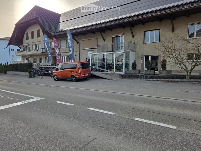 A two-story building with solar panels on the roof, a glass-enclosed entrance, and bicycles parked outside. The building has multiple windows and a balcony on the second floor. A red van is parked in front of the building.