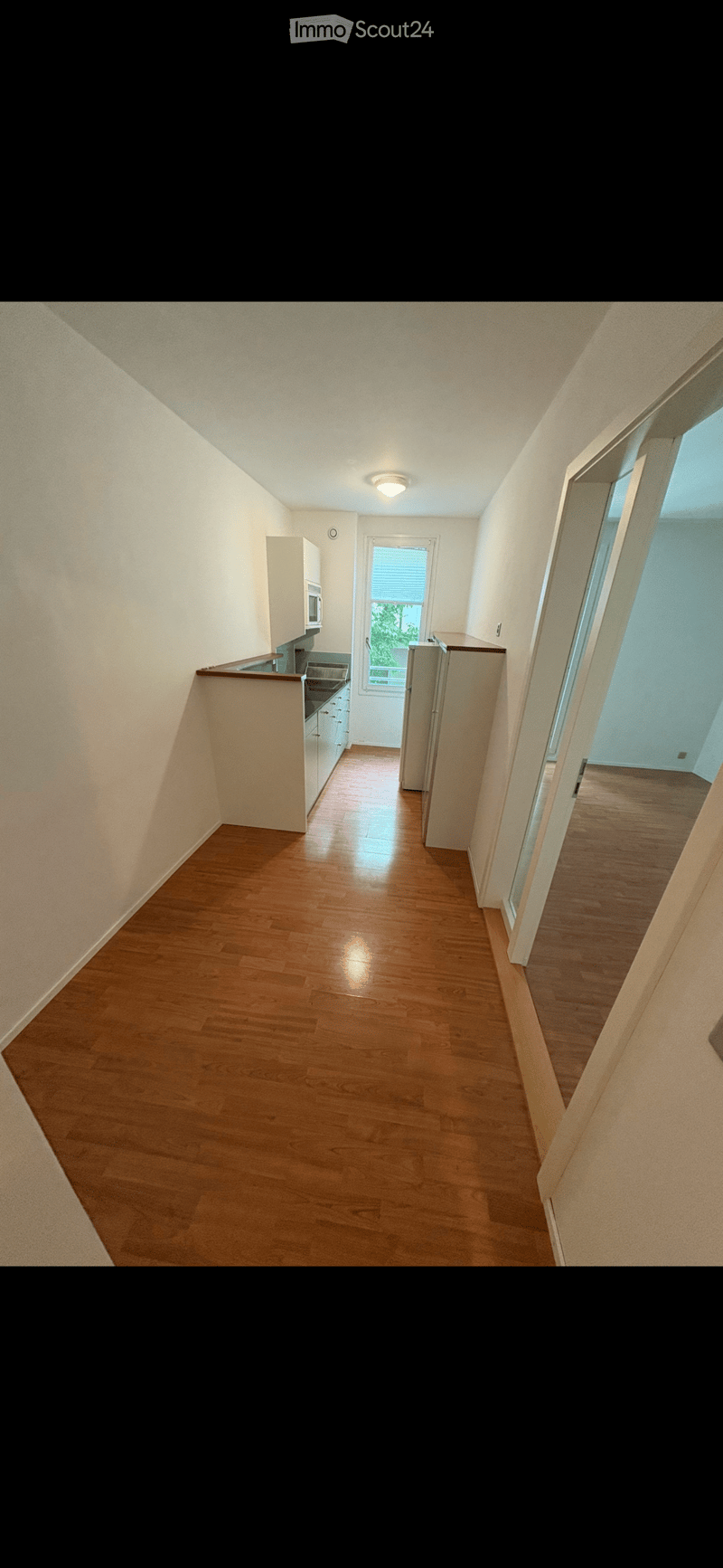 Empty room with wooden floor, white walls, kitchen area with sink and microwave, window with blinds, and a door leading to another room