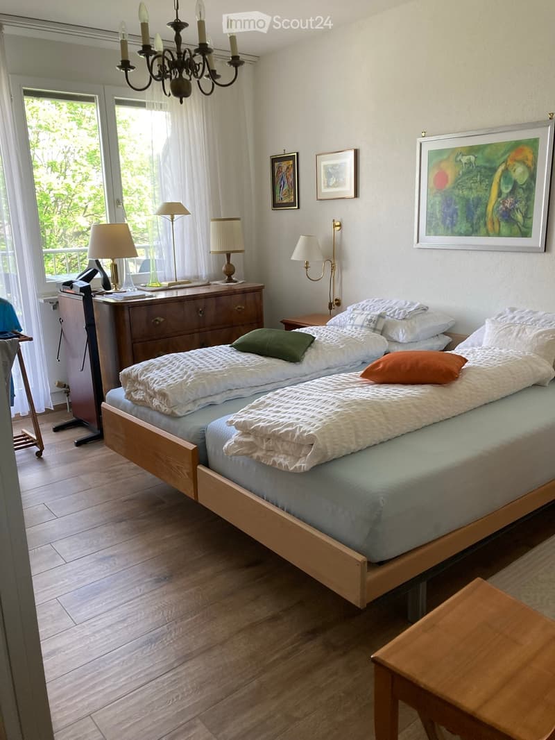 Two single beds, wooden floor, white walls, chandelier, lamps, wooden cabinet, paintings, telephone, window with curtains