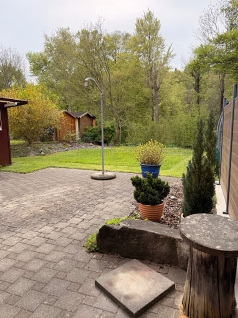 brick paved patio, wooden table, potted plants, lamp, wooden shed, grassy lawn, trees