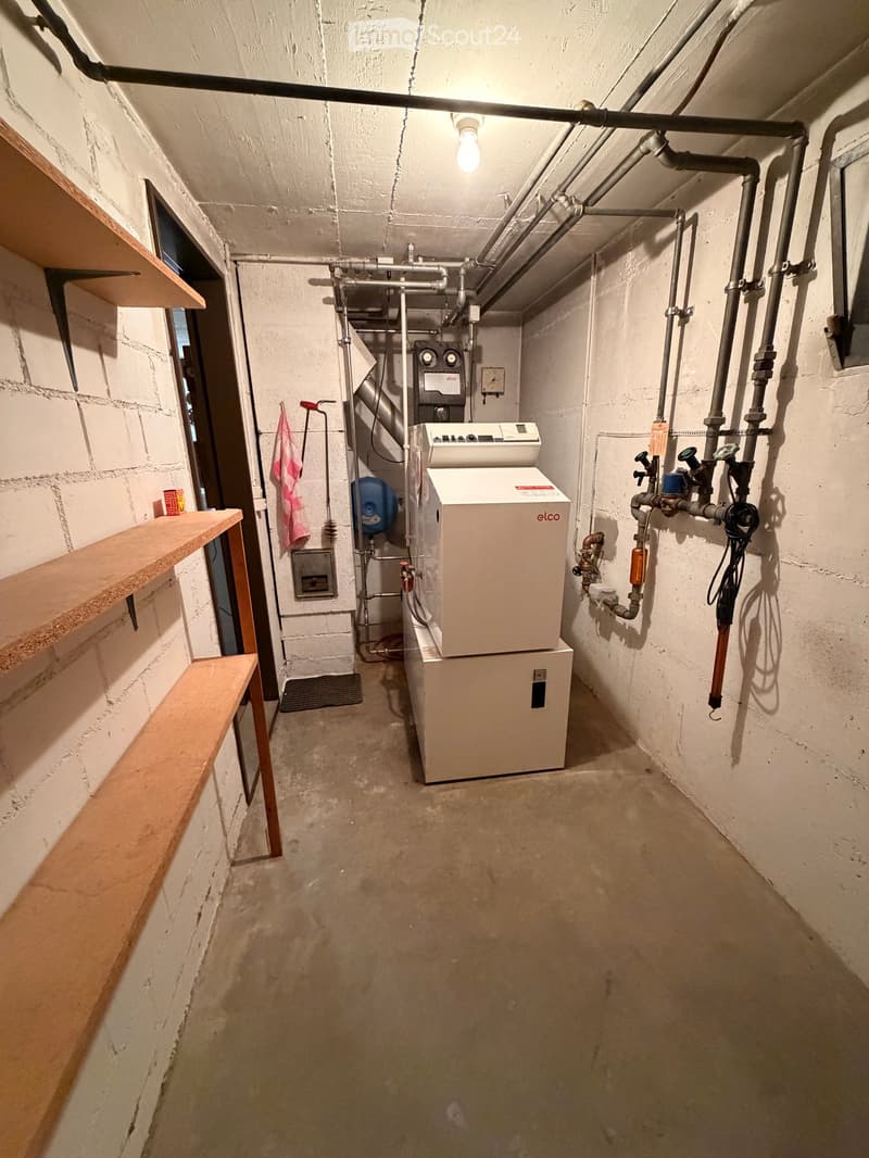 A basement room with white walls, concrete floor, wooden shelves, and two white appliances, one labeled 'elco' and the other a dishwasher.