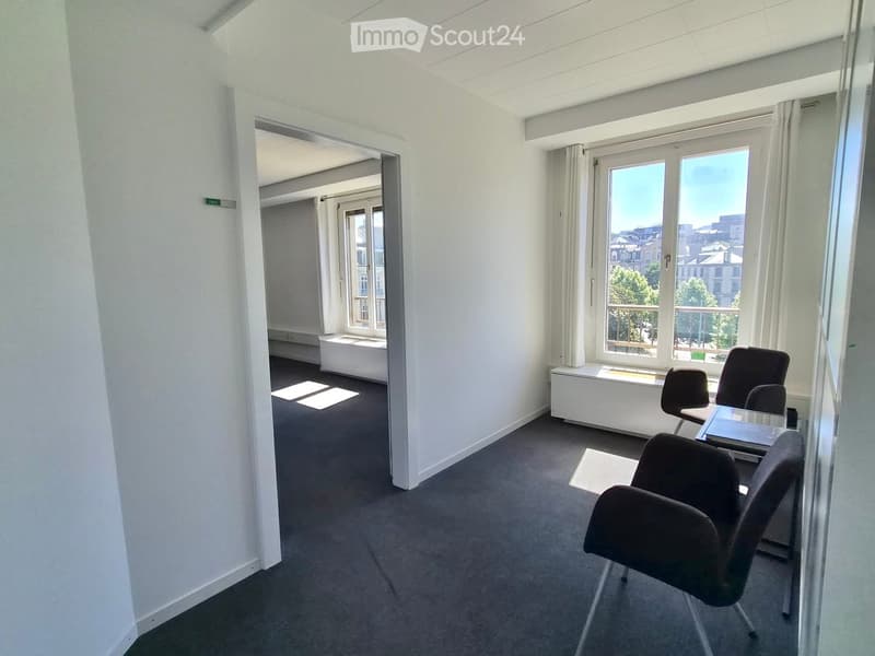 Office space with white walls, grey carpet, large windows with white curtains, and a view of the city. Includes a glass table and several black office chairs.