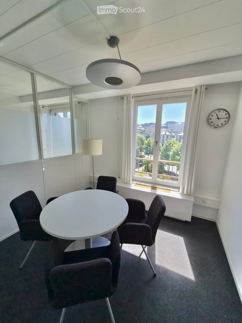 Modern furnished room with a round white table, four dark chairs, a floor lamp, a wall clock, and large windows with a view of the city.