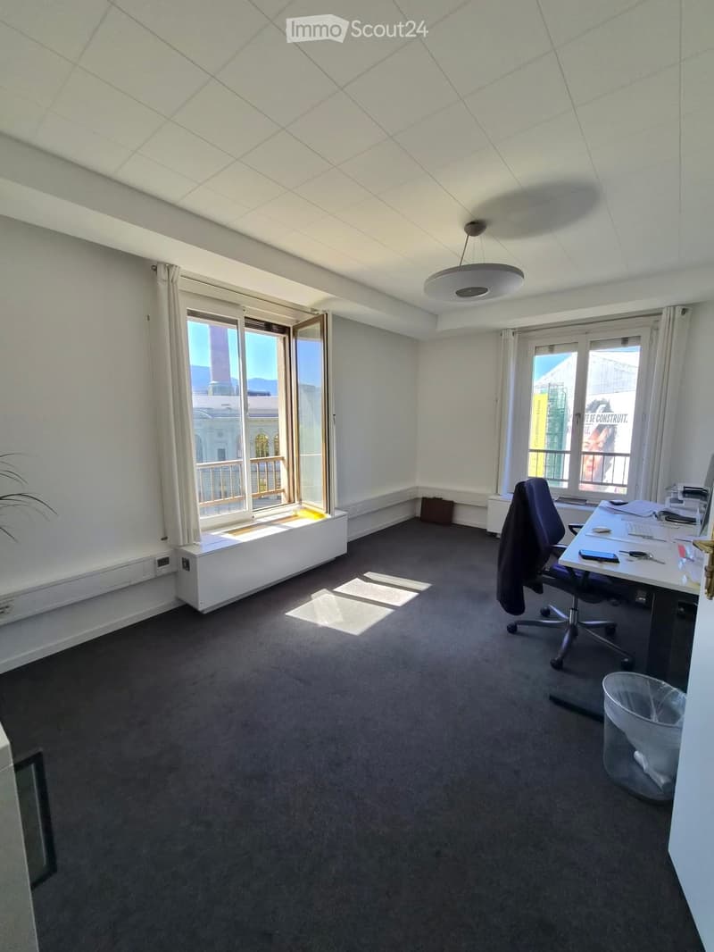 Office space with a desk, chair, computer screen, and trash bin. Large windows with white curtains and a view of the city.