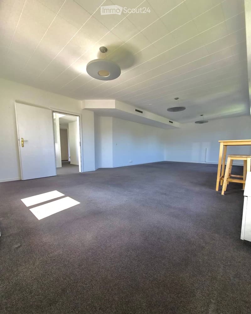 Empty room with grey carpet, white walls, and a ceiling light fixture. A door is visible on the left, and a small table and chair are placed in the corner on the right.