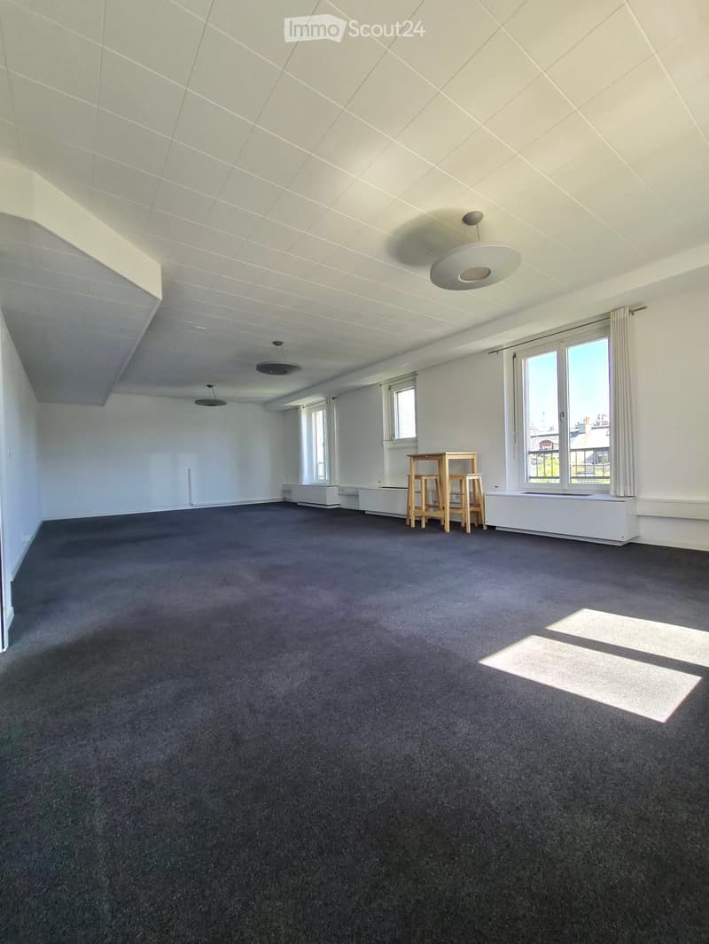 Empty room with grey carpet, white walls, large windows, and a small wooden table with two chairs in the corner.