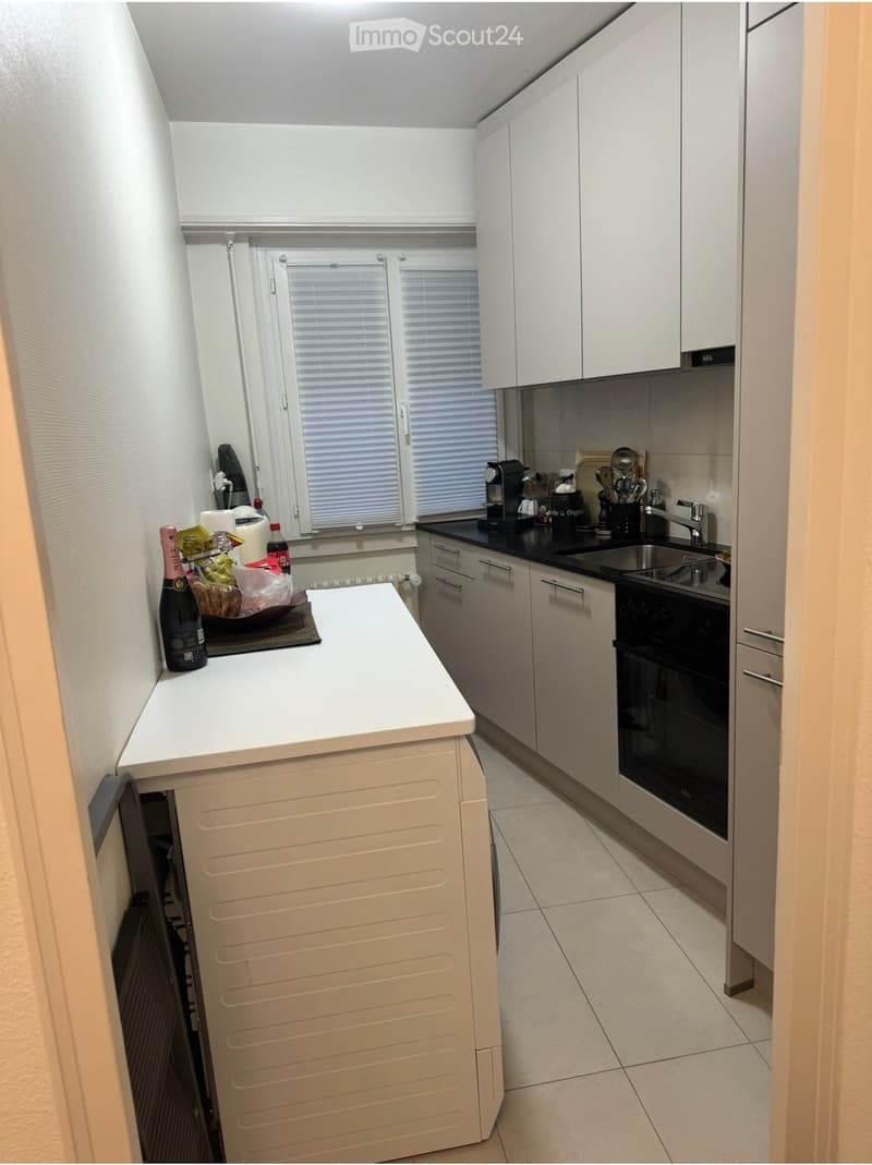 modern kitchen with white cabinets, black countertops, white tiled floor, white island with a bottle, sink, oven, dishwasher, and a window with blinds