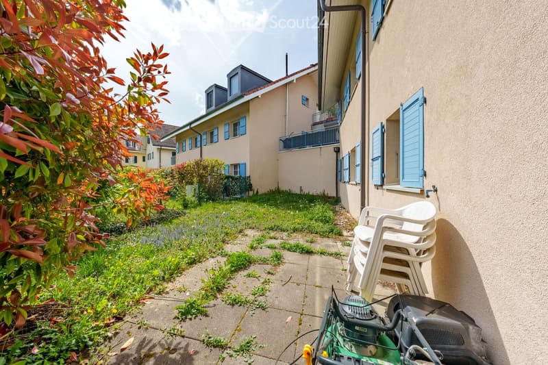 A beige building with blue shutters, a small garden, and a lawn mower