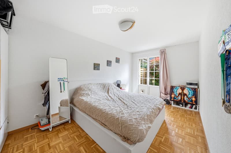 white bed, white headboard, white bedspread, mirror, wooden floor, white walls, window with curtains, small table, lamp, shelf with books and decorative items