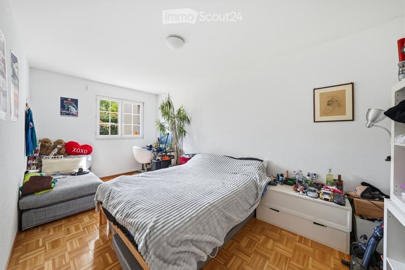 Bedroom with bed, desk, chair, plant, window, and various items on the floor and shelves.
