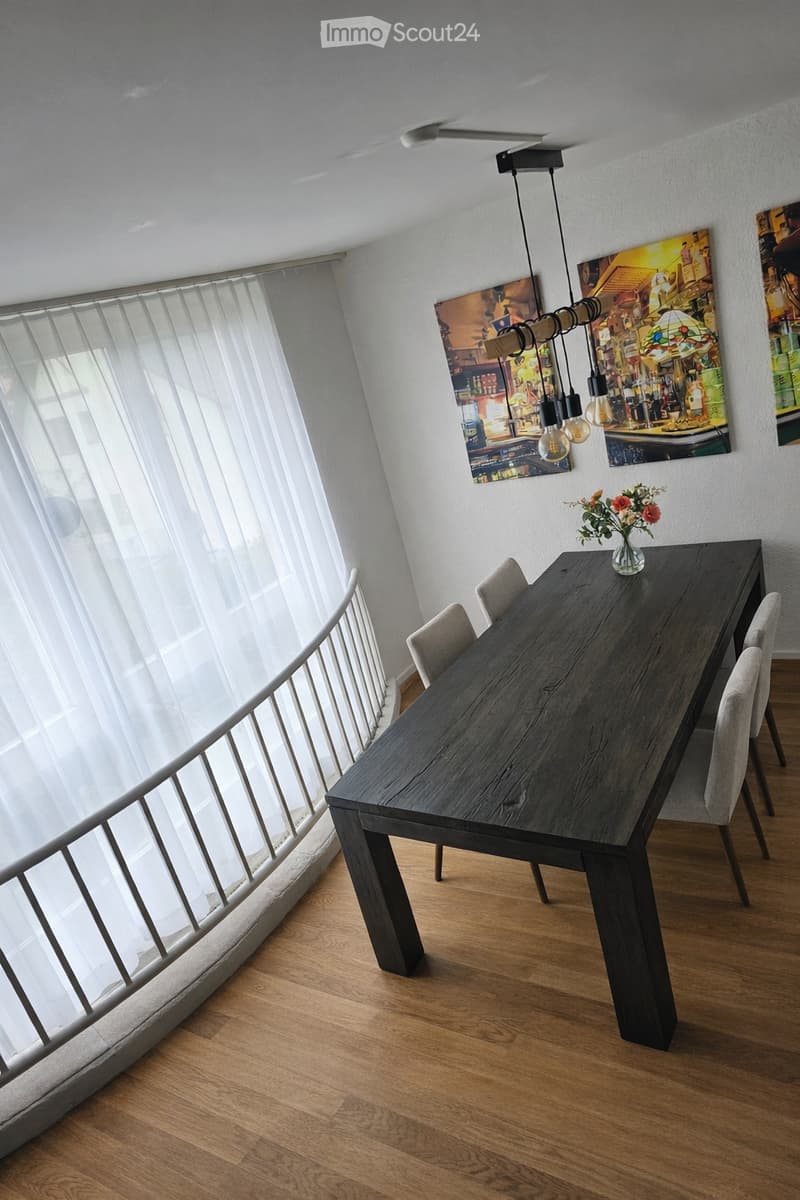 Dining room with a large wooden table, 4 chairs, a flower vase, and a chandelier. The room has a white wall with three paintings, a white curtain, and a balcony with a metal railing.