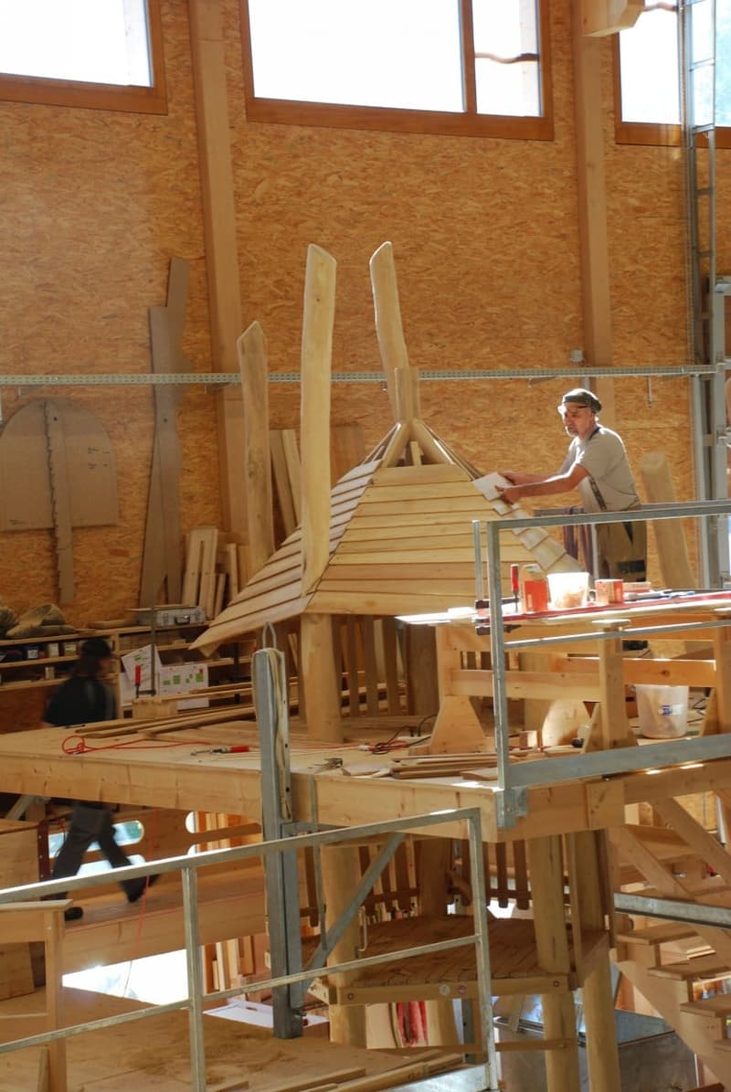 Wooden structure being constructed, wooden walls, large wooden beams, multiple work tables, and ladders.