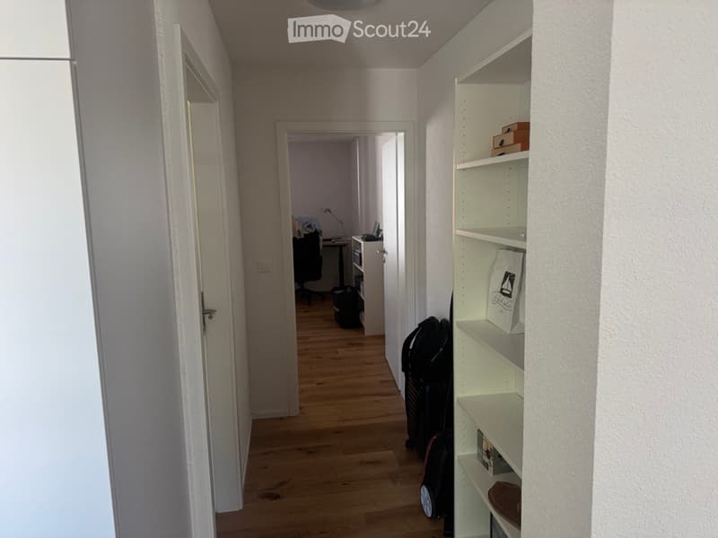 Empty hallway with wooden floor, white walls, white shelves, and a door leading to another room with a desk and chair.