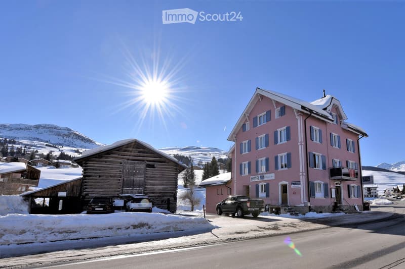 Two buildings, one pink and one wooden, with cars parked in front, snowy mountains in the background