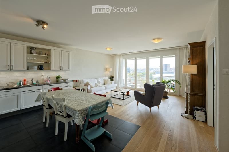 Open plan living room and dining area with large windows, white kitchen, dining table, couch, armchair, coffee table, floor lamp, rug, potted plant