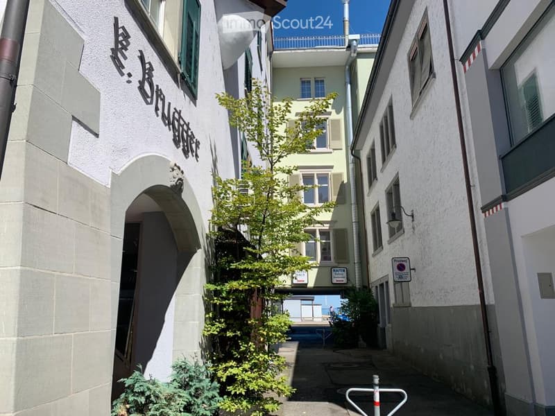 Narrow alley between two buildings, one with a sign in a foreign language, the other with a balcony and windows, a tree in the middle, and a metal barrier at the end.