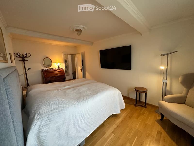 A bedroom with a bed, a wooden cabinet, a mirror, a lamp, a TV, a chair, a stool, and a coat rack.