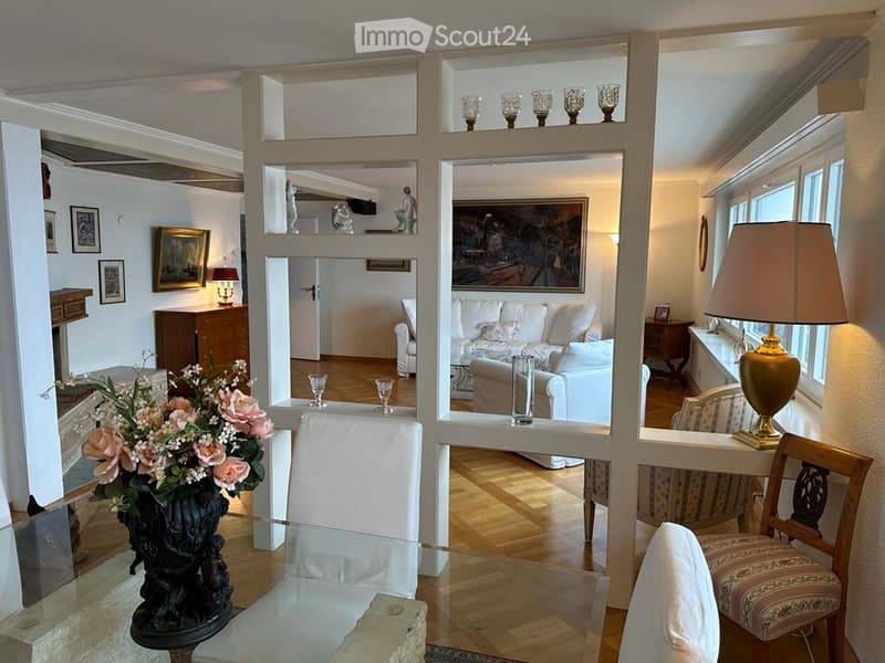 Dining room with glass table, white chairs, white couch, wooden floor, paintings, lamps, flower vase