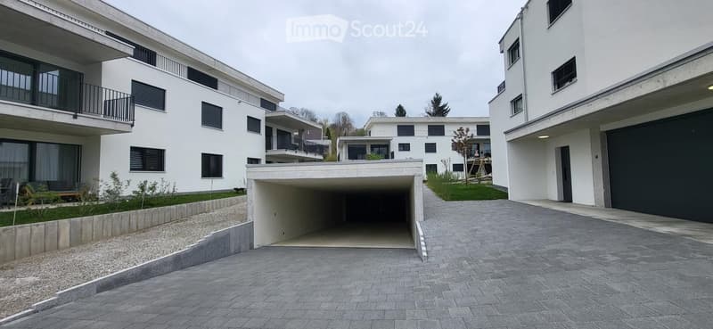 Modern apartment complex, white exterior, underground garage, paved driveway, balconies, greenery