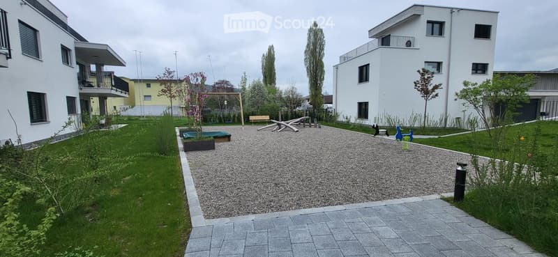 A gravel path leads to a small playground with a swing and a bench. The garden is surrounded by grass and has a small tree and a planter.