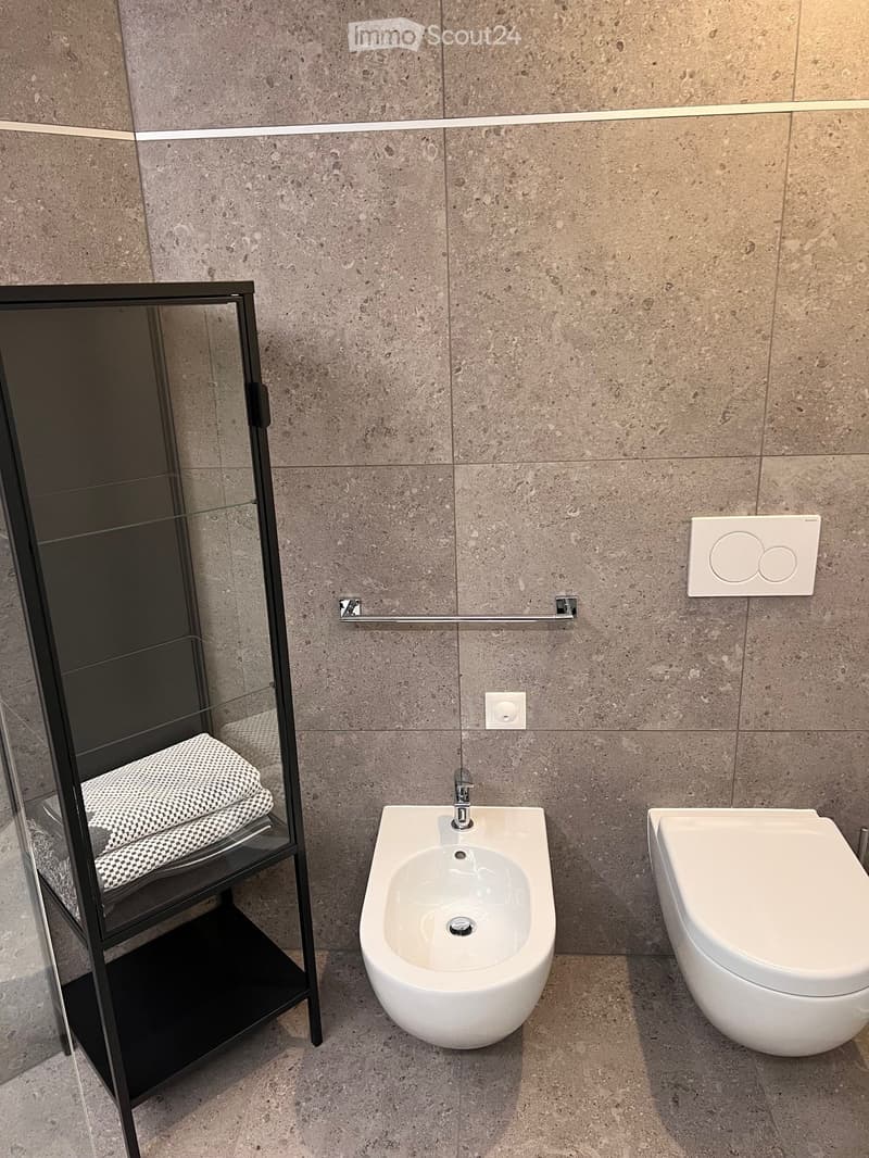 Modern bathroom with white fixtures, gray tiled walls, bidet, toilet, and a glass shelf with towels.
