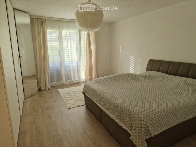Bedroom with a wooden floor, large window with blinds and curtains, a bed with a patterned duvet, a mirror, and a small white rug.