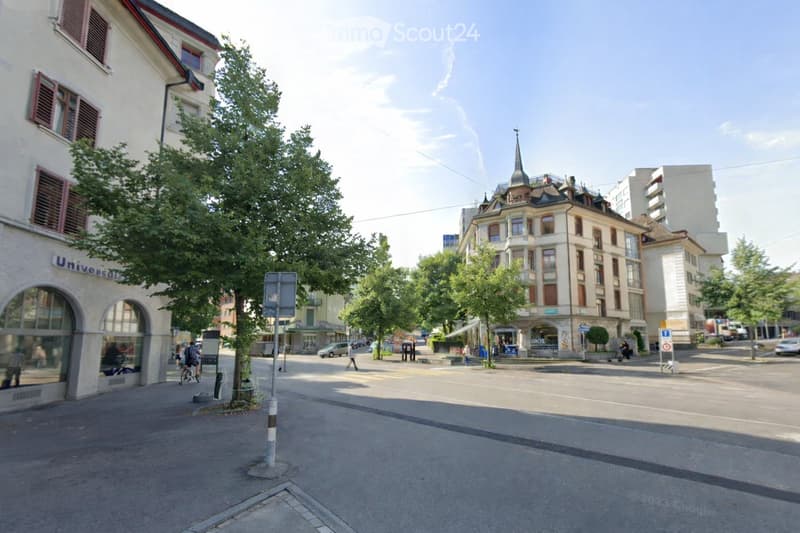 A street with buildings on both sides, a tree in the middle, and people walking on the sidewalk.
